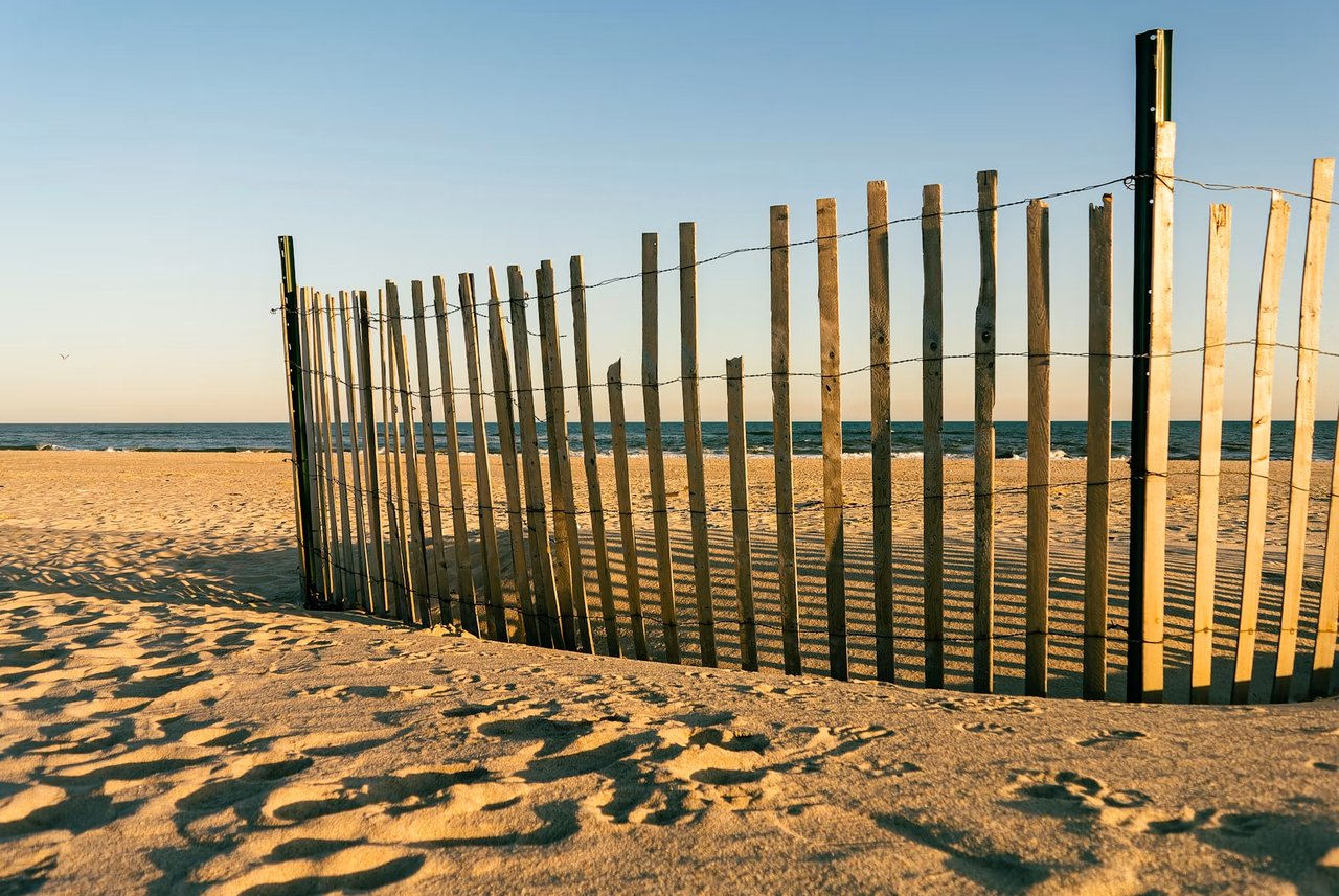 A wooden beach fence casts long shadows across the sand, with the ocean in the background.