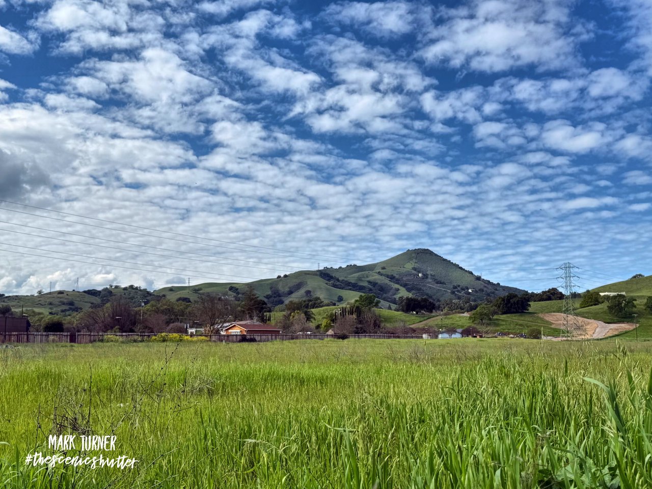 Green field and rolling hills beneath a mountain ridge and cloud-filled sky.