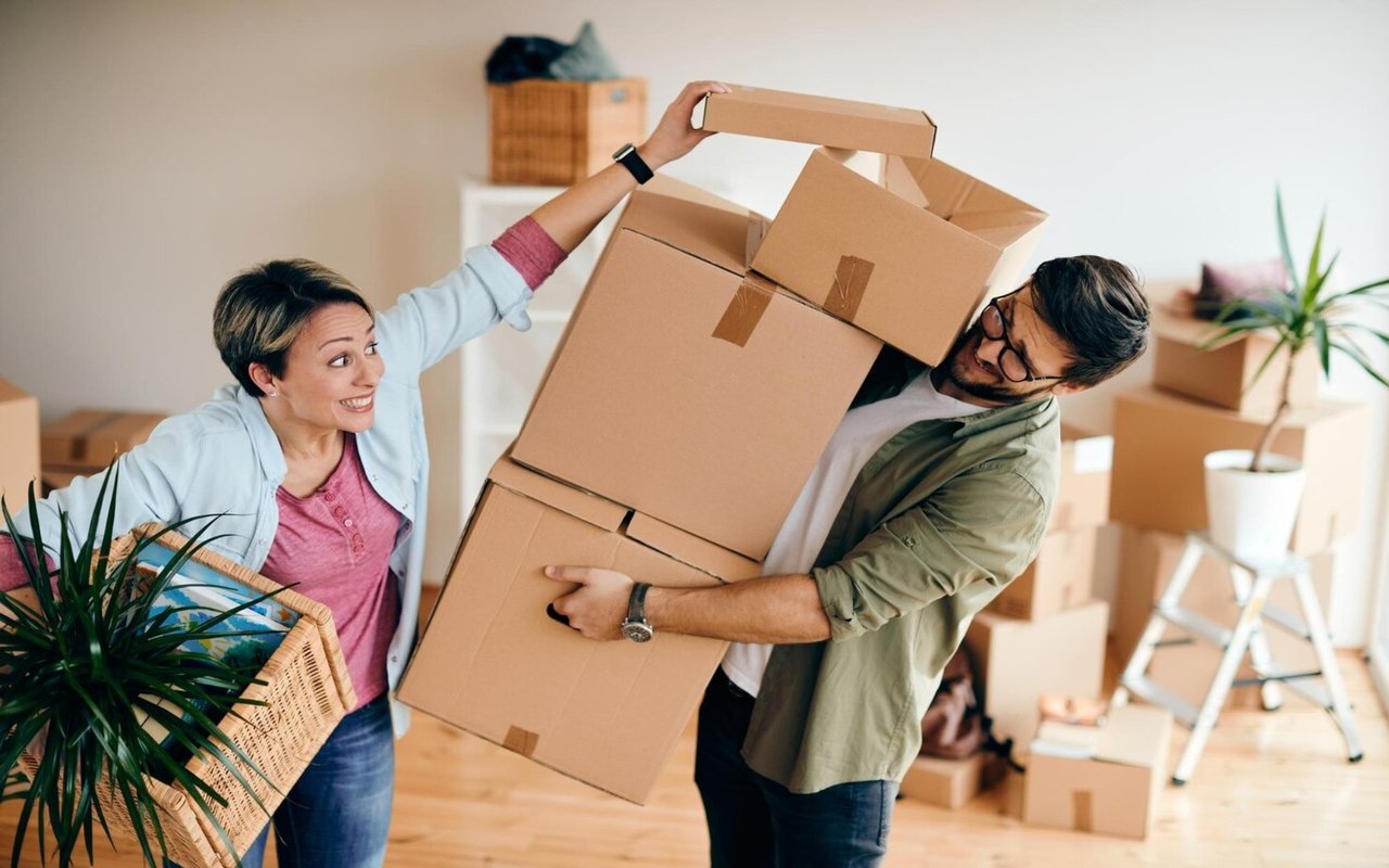 A couple carries stacked moving boxes through a bright home.