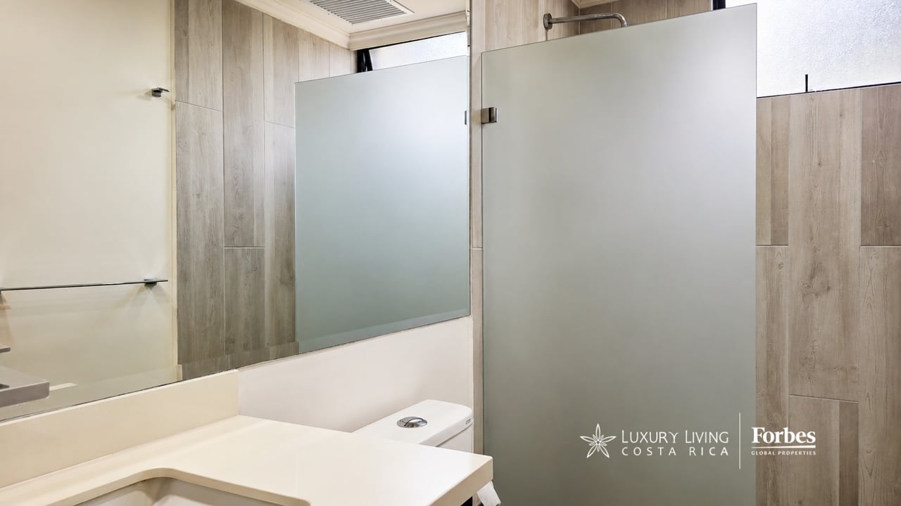 Modern bathroom with a white vanity, large mirror, wood-look tile, and frosted-glass shower enclosure.