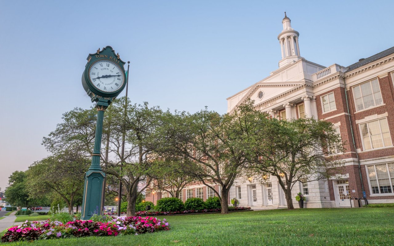 Greenwich Town Hall behind leafy trees, with a green street clock and flower beds in front.