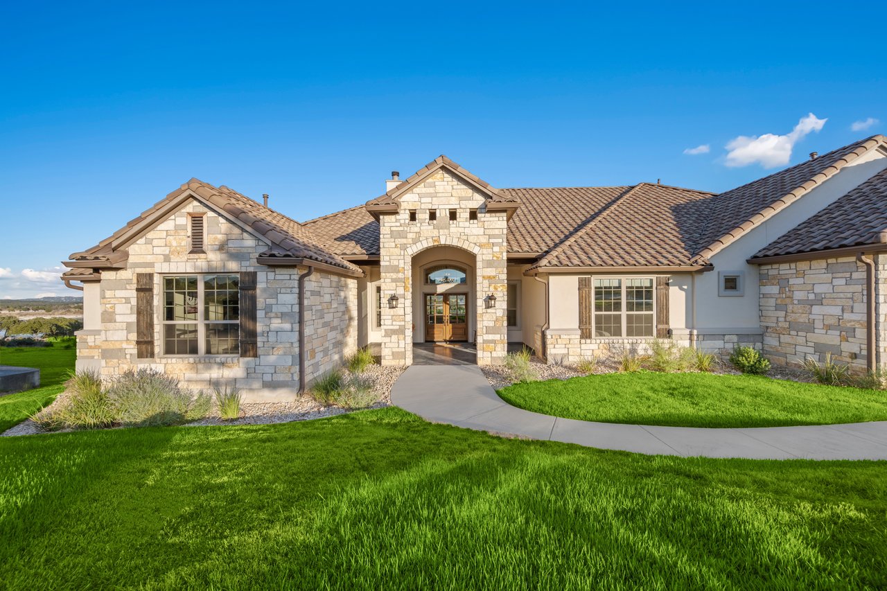 Stone-and-stucco house with a tiled roof, arched entry, curved walkway, and broad green lawn.