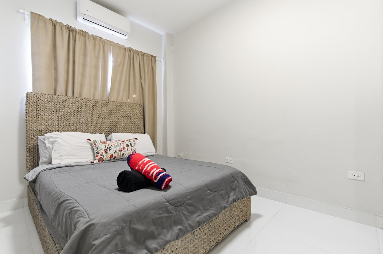 Bedroom with a gray-covered bed, woven rattan headboard, beige curtains, and wall-mounted air conditioner.