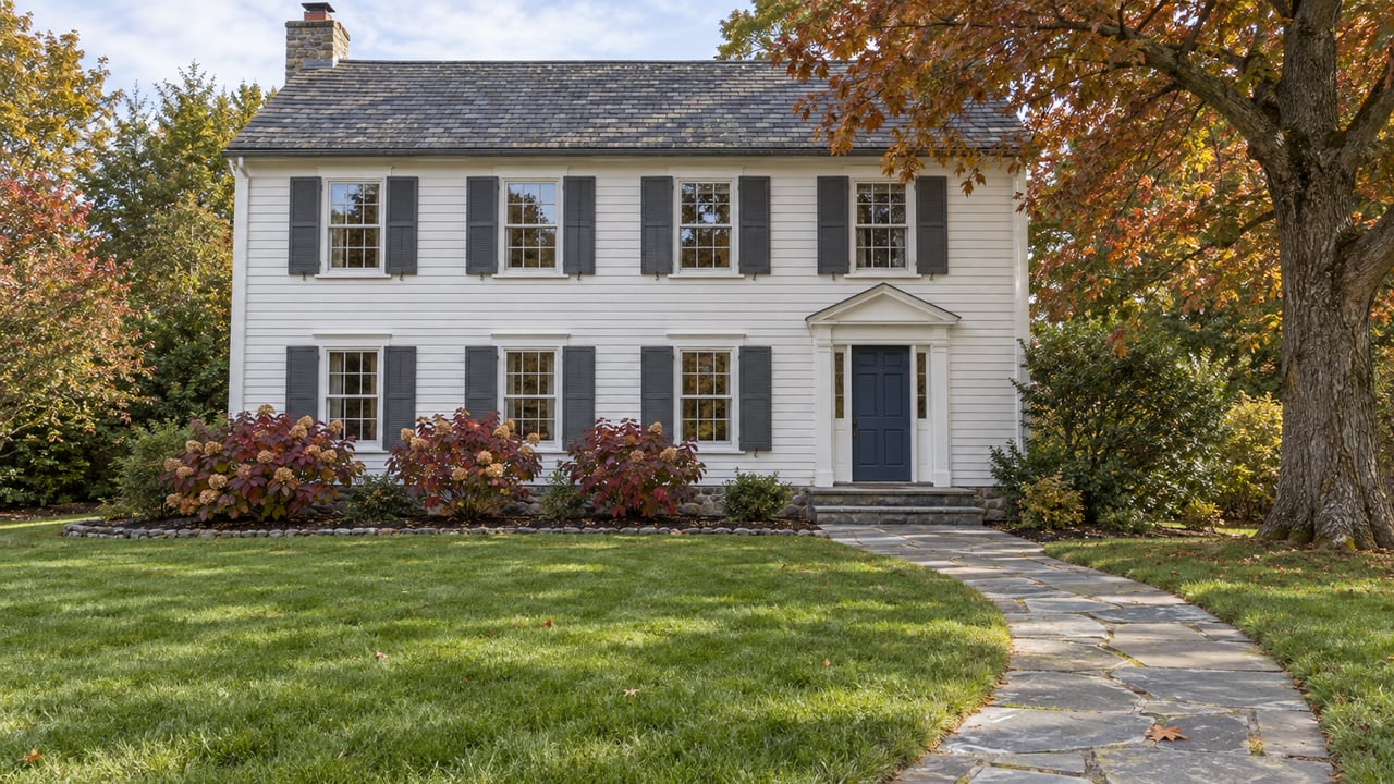 An established white clapboard home with a slate roof, dark shutters, and a blue front door beyond a broad lawn and bluestone walk, framed by an oak in autumn color.