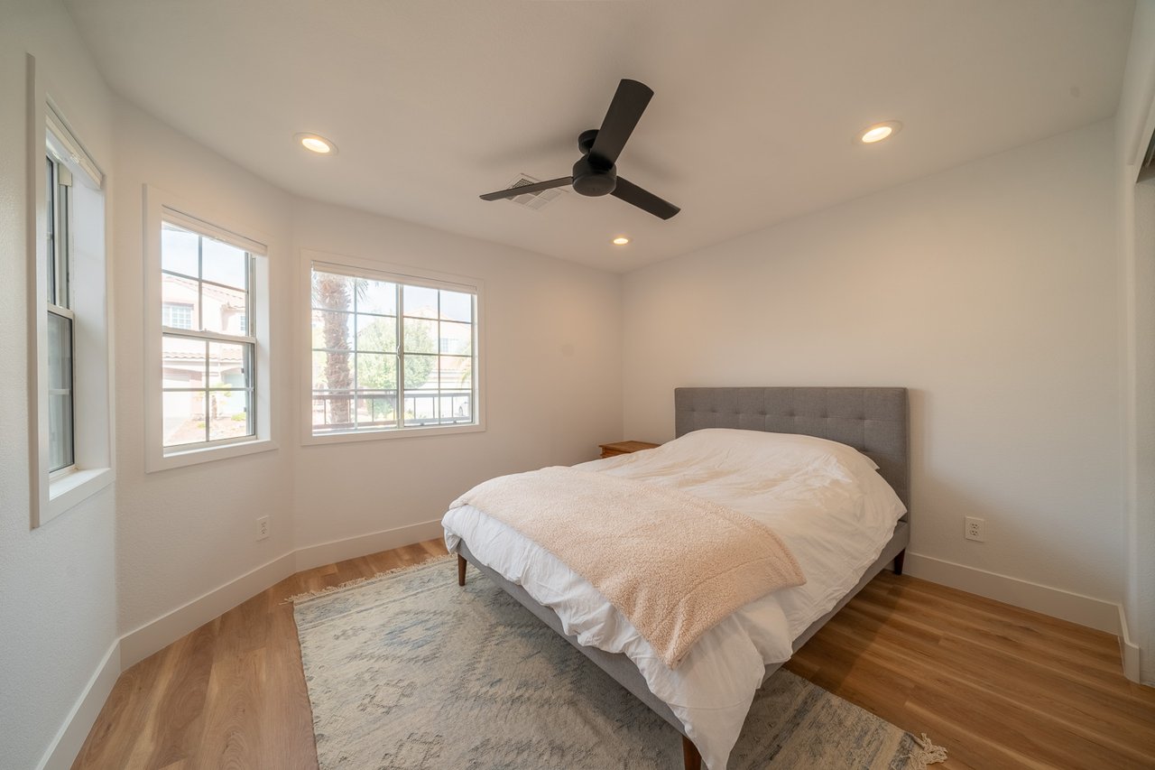 Bedroom with a gray upholstered bed, three windows, a black ceiling fan, and wood floors.