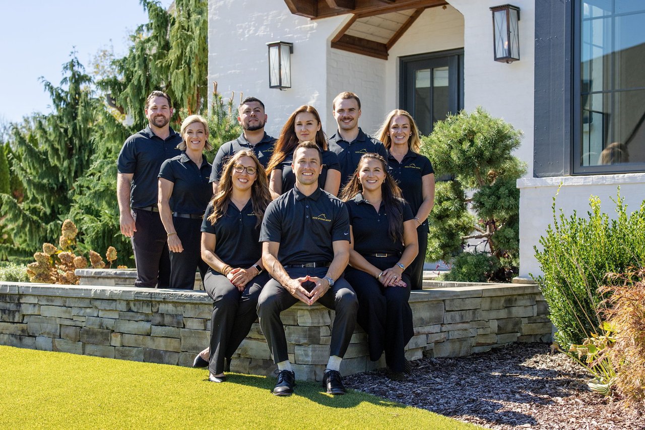 Nine real estate agents pose outside a white home, three seated in front of six standing teammates.