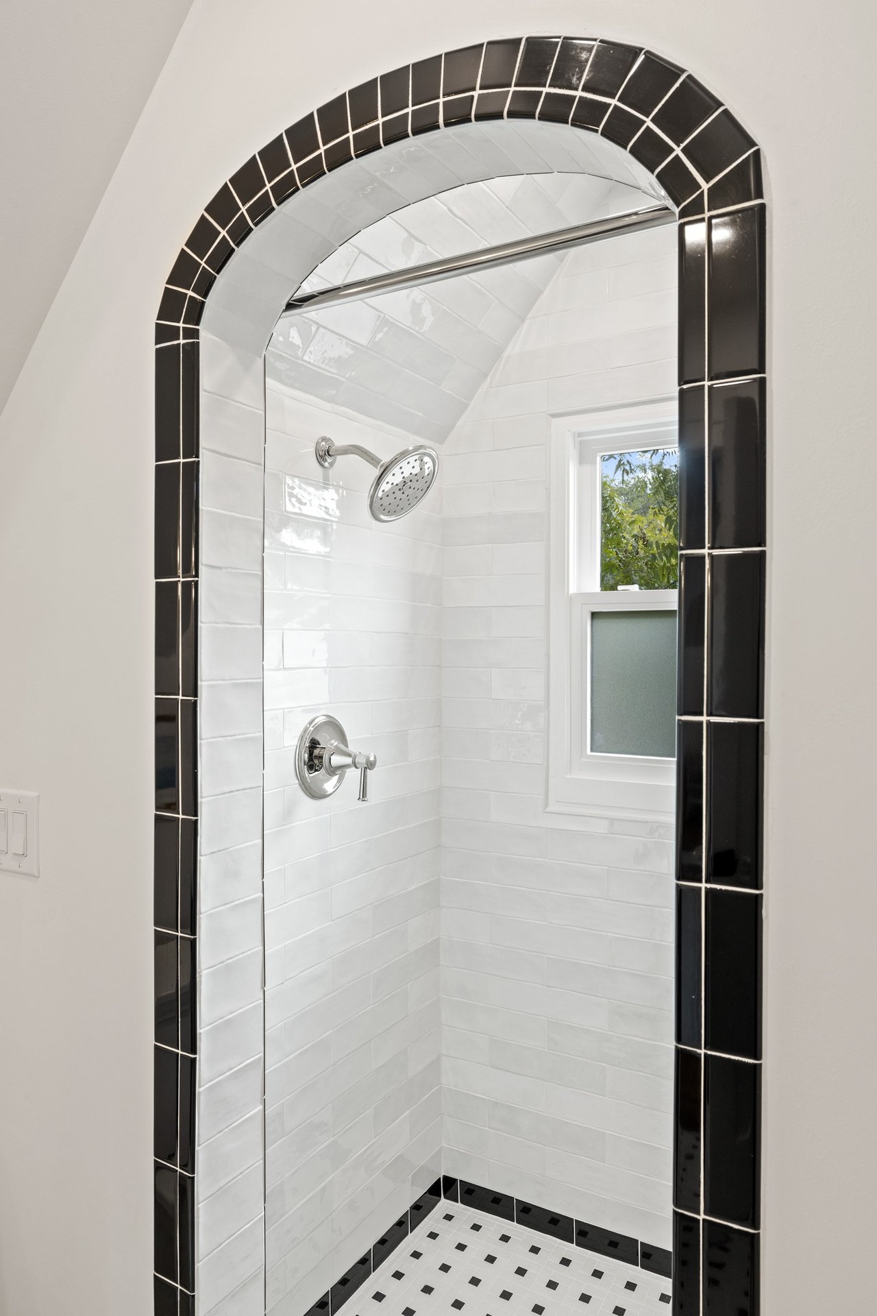 Tiled shower with an arched black-tile surround, white walls, chrome fixtures, and a small window.