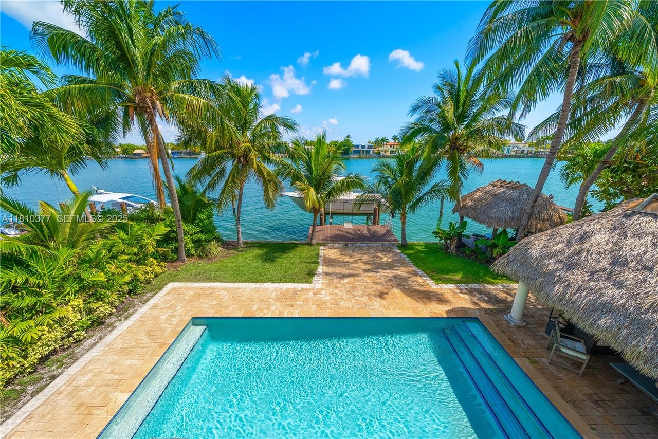 Waterfront pool and patio framed by palm trees, with a dock and thatched tiki huts.