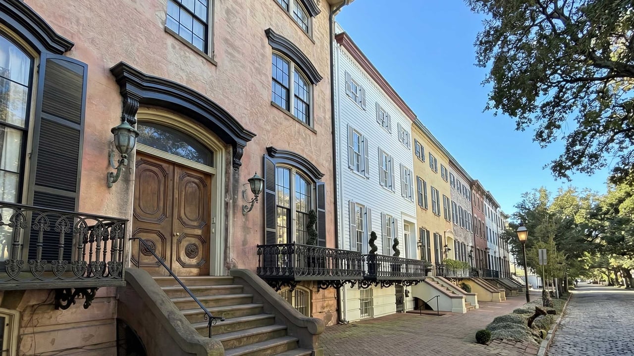 A side-angle view of a historic Savannah townhome featuring a high brownstone stoop, ornate black wrought iron railings, and a grand arched wooden double door against a clear blue sky.