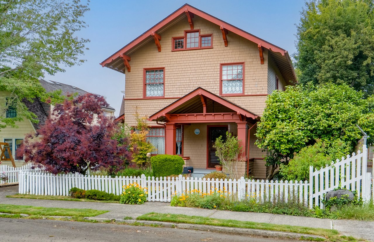 Front exterior of a two-story Craftsman-style home with tan cedar-shingle siding, red trim, a covered front porch, mature landscaping, flowering shrubs, and a white picket fence along a quiet residential street.