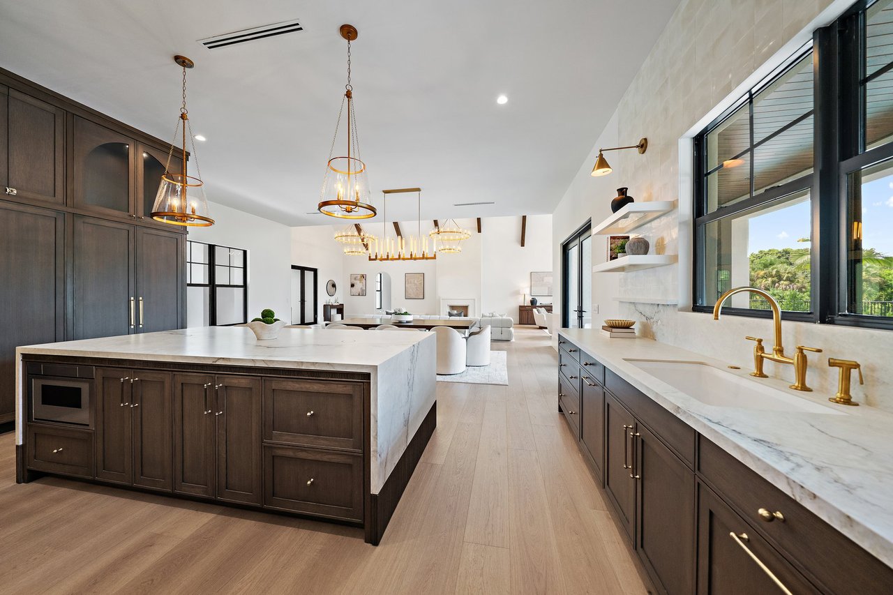 Modern kitchen with a large marble island, dark cabinetry, gold fixtures, and tall windows.