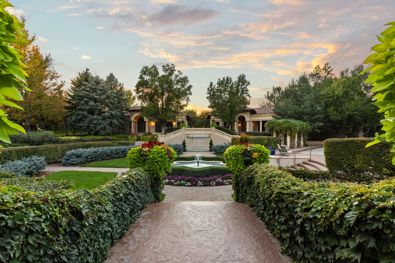 Formal estate garden with a fountain, small bridge, flower beds, and a stone path at sunset.