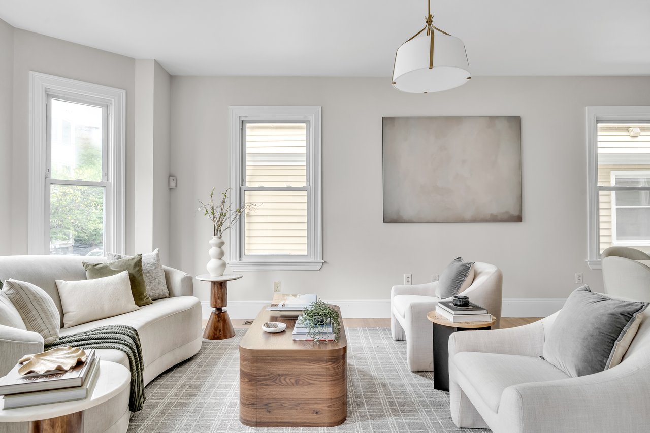 Bright staged living room with cream seating, a wood coffee table, and tall windows.