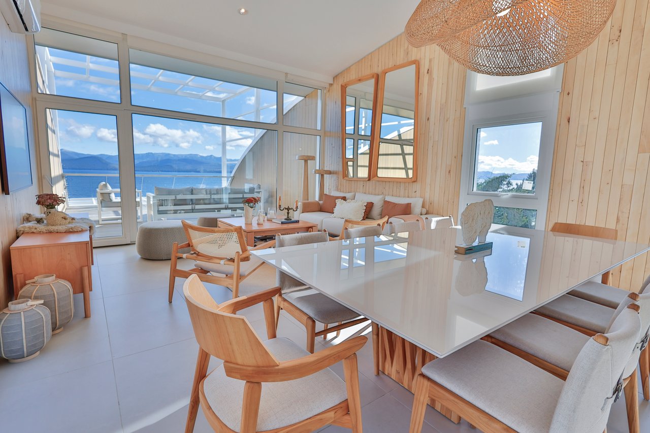 Dining and living area with pale wood furniture and large windows overlooking a lake and mountains.