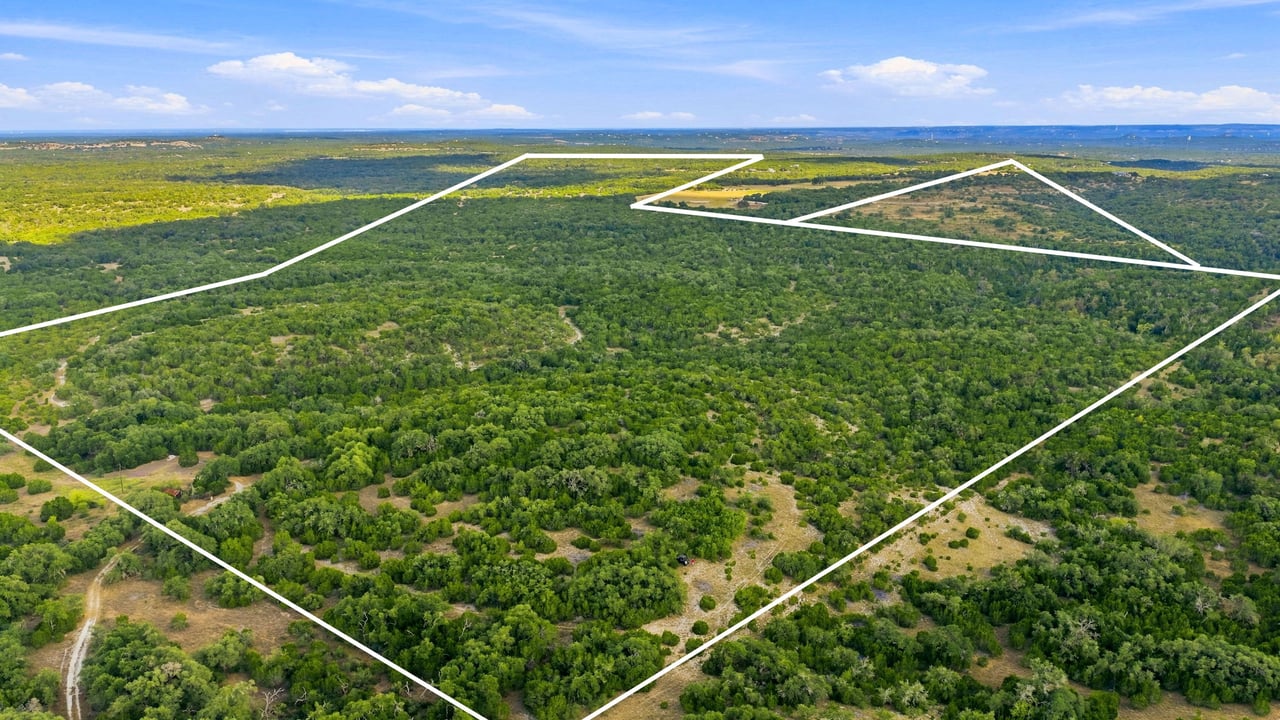 Aerial view of wooded ranch acreage outlined by white property boundaries, with open parcels in the distance.