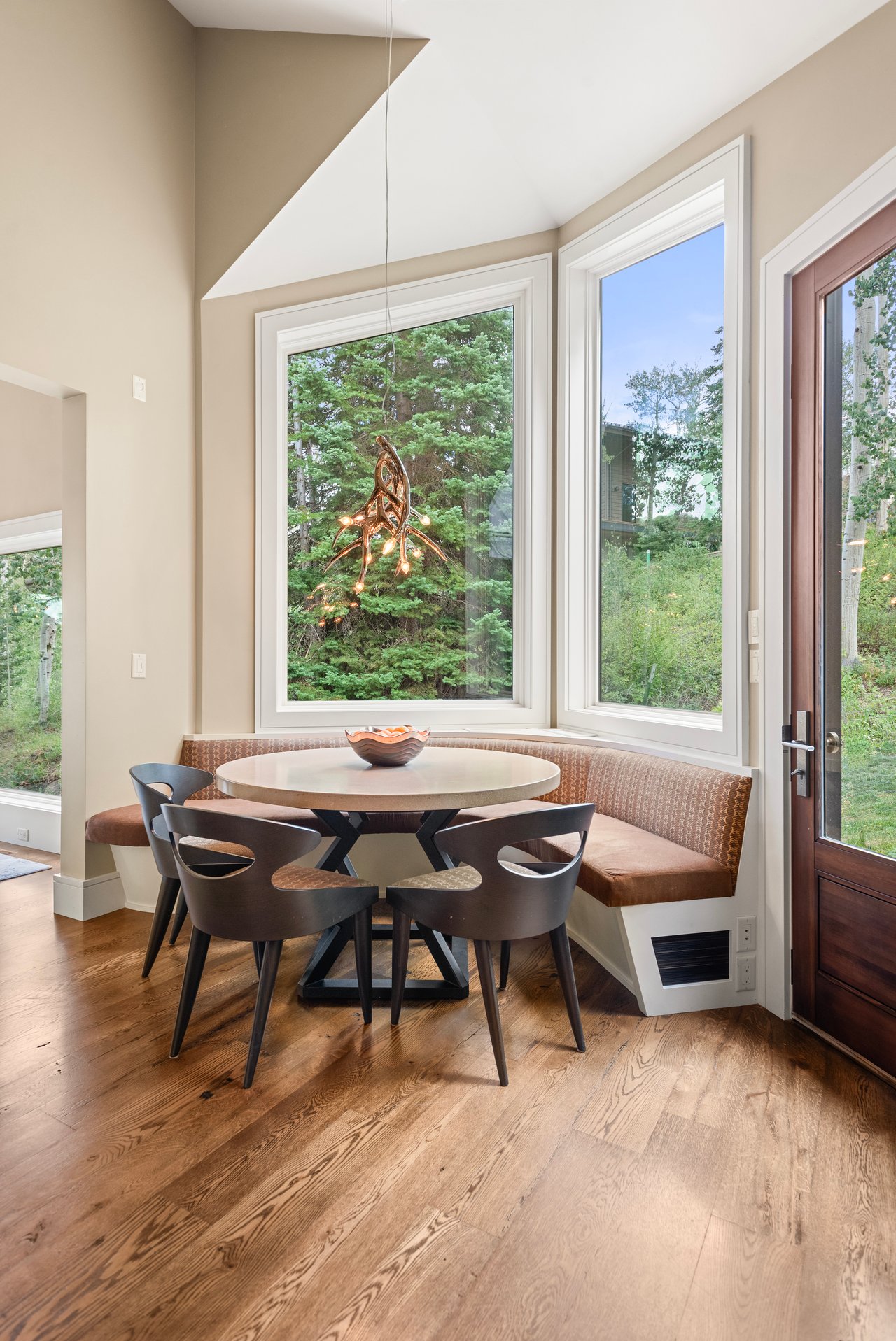 Breakfast nook with a round table, curved bench, and tall windows overlooking trees.