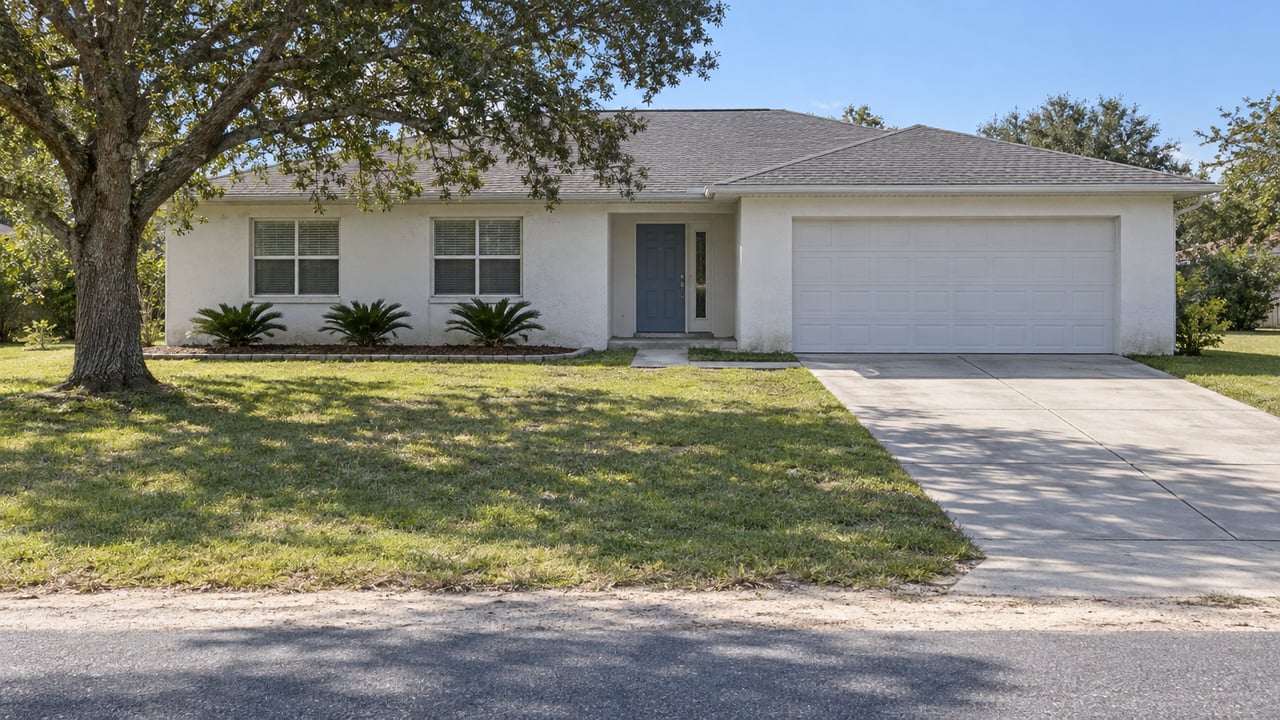 Light-stucco single-story house behind a deep front lawn, with a spreading live oak and a concrete driveway in bright afternoon light.