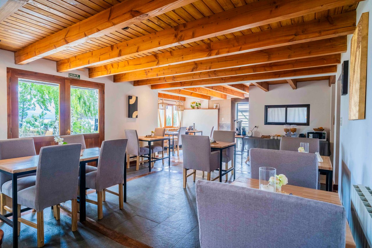 Hotel dining room with exposed timber ceiling, upholstered chairs, large windows, and a buffet counter.