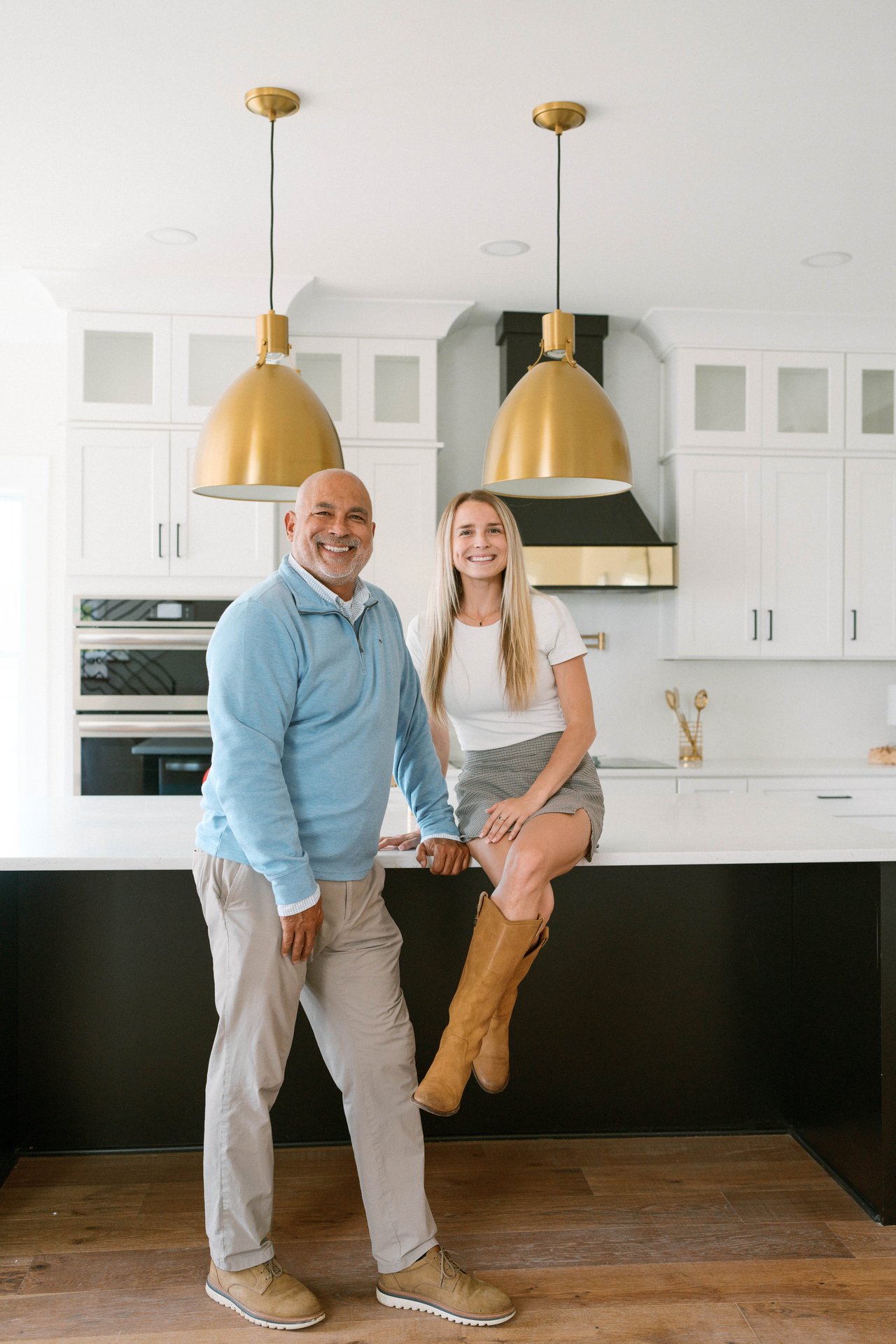 Two smiling adults pose in a bright kitchen beneath brass pendant lights.
