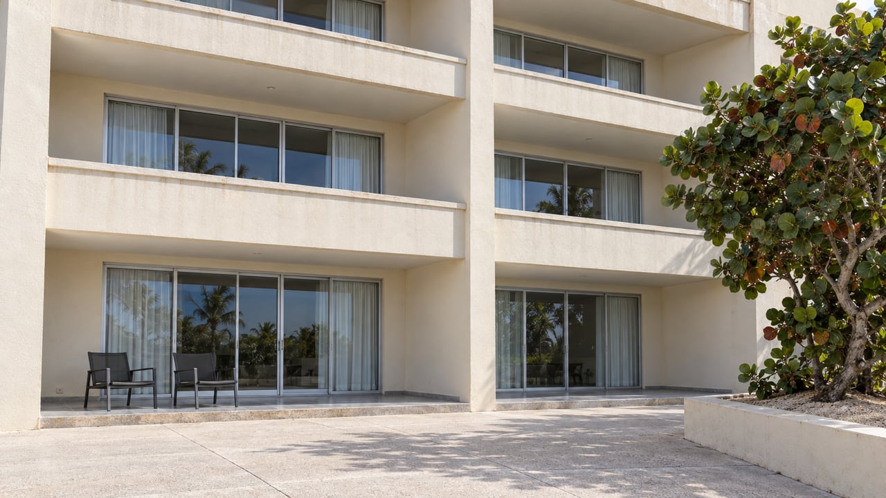 Cream stucco condominium balconies and closed glass doors above a pale concrete courtyard, with a mature sea grape in a raised masonry bed.