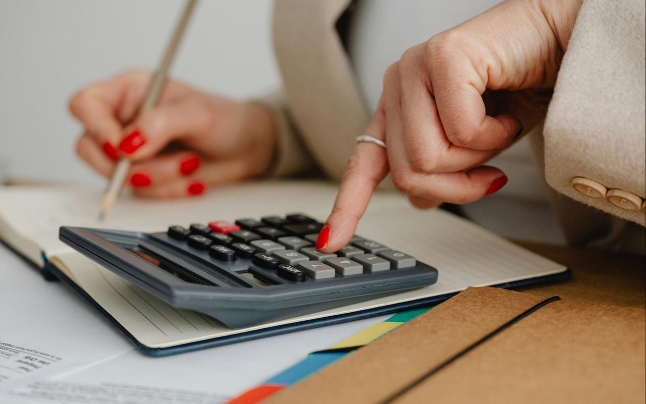 Hands use a calculator and write notes on paperwork beside a brown folder.