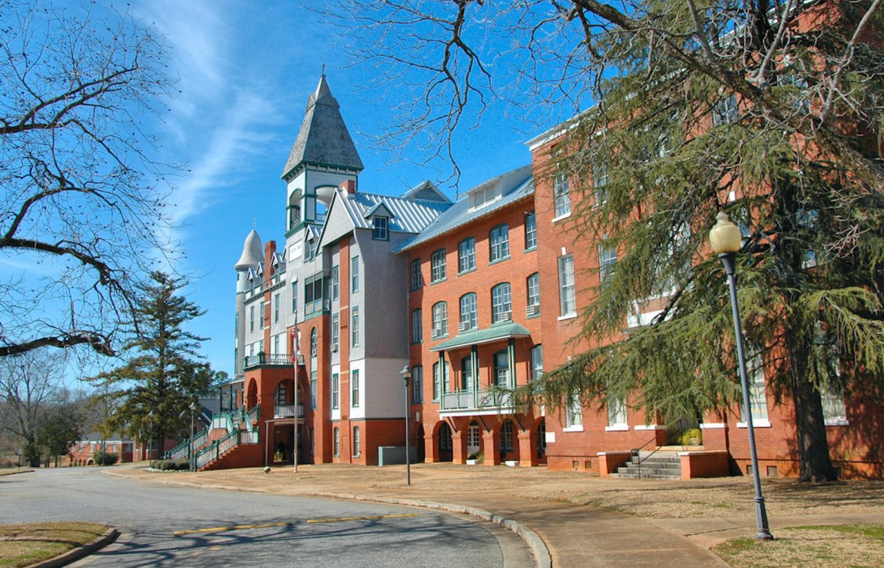 Multi-story brick building with a pointed tower beside a quiet, tree-lined street.