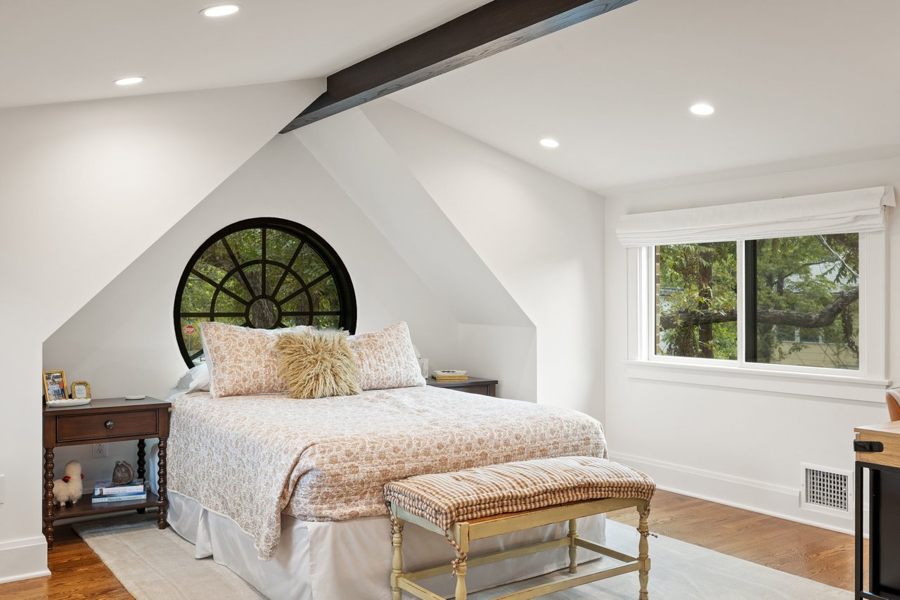 Bedroom with a round black-framed window behind the bed and a dark exposed ceiling beam.