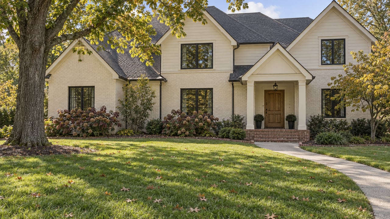 A two-story painted-brick home behind a deep lawn, with black-framed windows, an oak tree and early autumn planting.