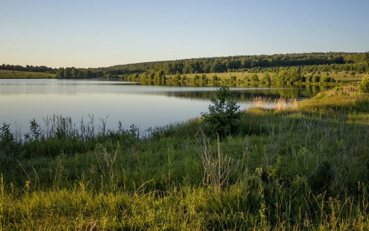 A calm lake bordered by grassy banks and wooded hills in warm evening light.