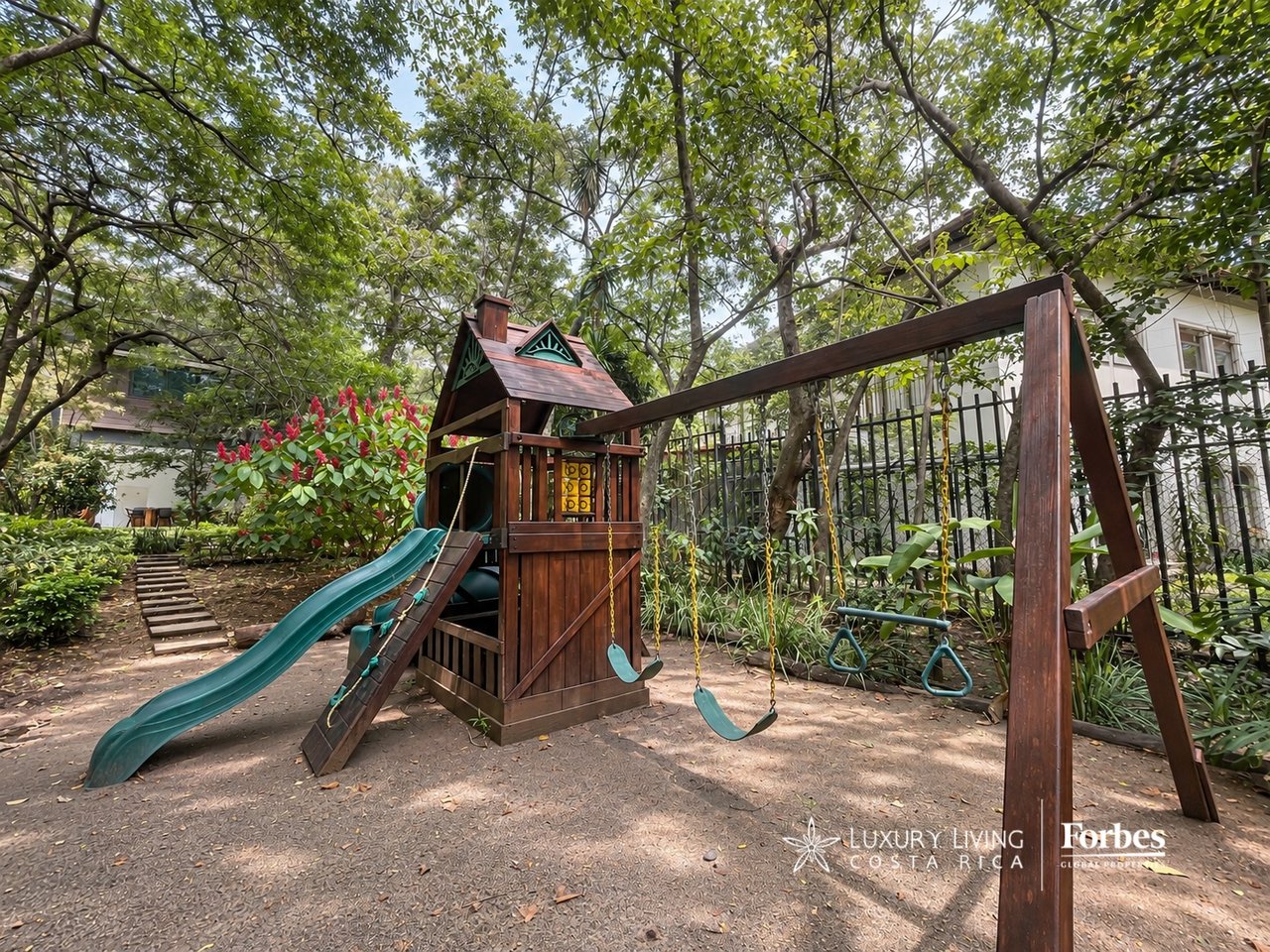 Outdoor playground with a wooden play structure, teal slides, swings, and mature trees.