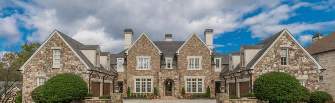 Large stone manor house with symmetrical gabled wings and a landscaped front entrance.
