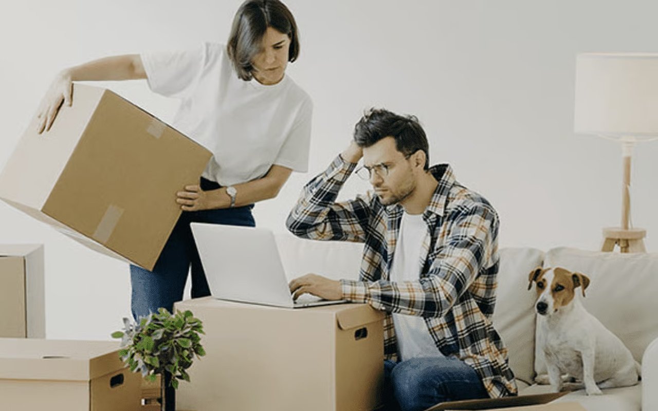 A man browses on a laptop among moving boxes as a woman and their dog sit nearby.