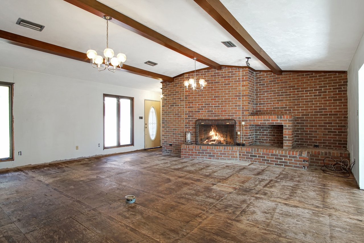 Spacious living room with a brick fireplace on the right, emitting a cozy fire. Wooden beams accent the ceiling, and light fixtures add warmth.