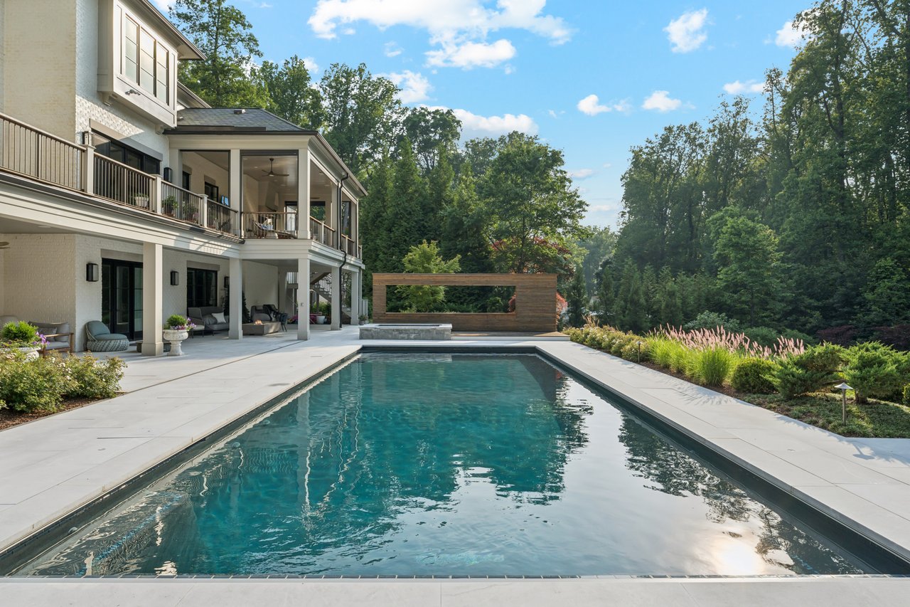 Backyard swimming pool beside a modern white home, with covered seating and wooded landscaping.