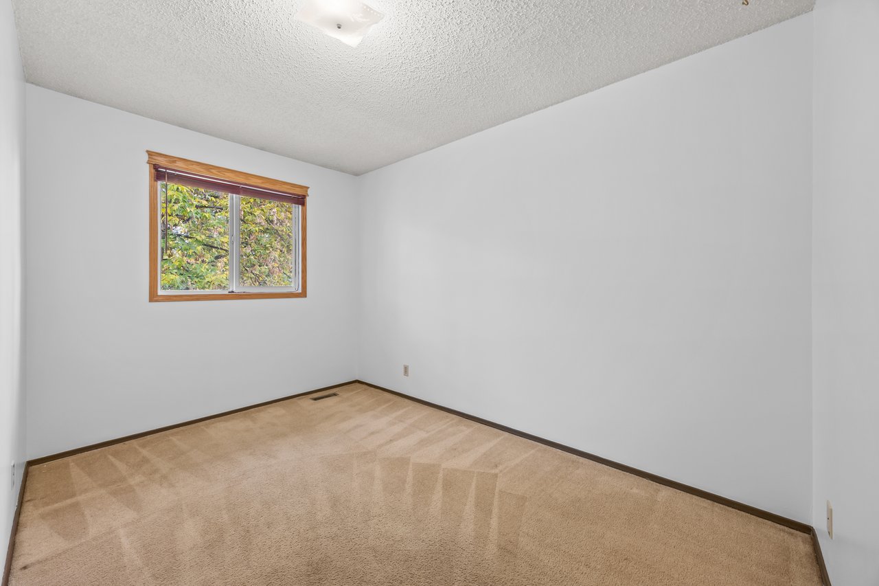 Empty bedroom with beige carpet and a wood-trimmed window overlooking leafy trees.