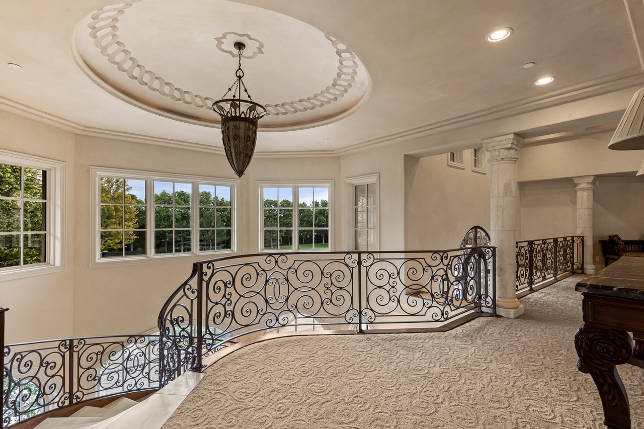 Upstairs landing with an ornate iron stair railing, patterned carpet, chandelier, and large windows.