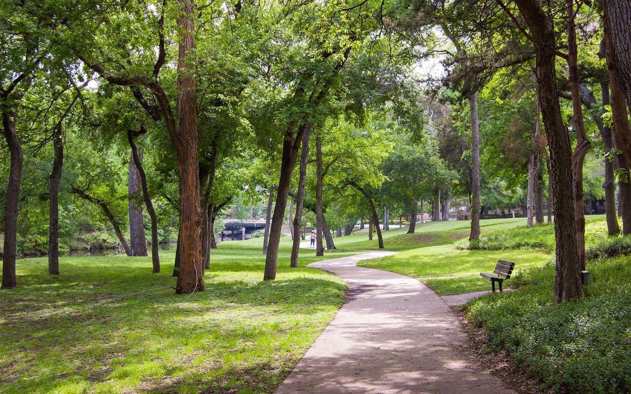 Curving park path beneath mature trees, with a bench and pedestrian in the distance.