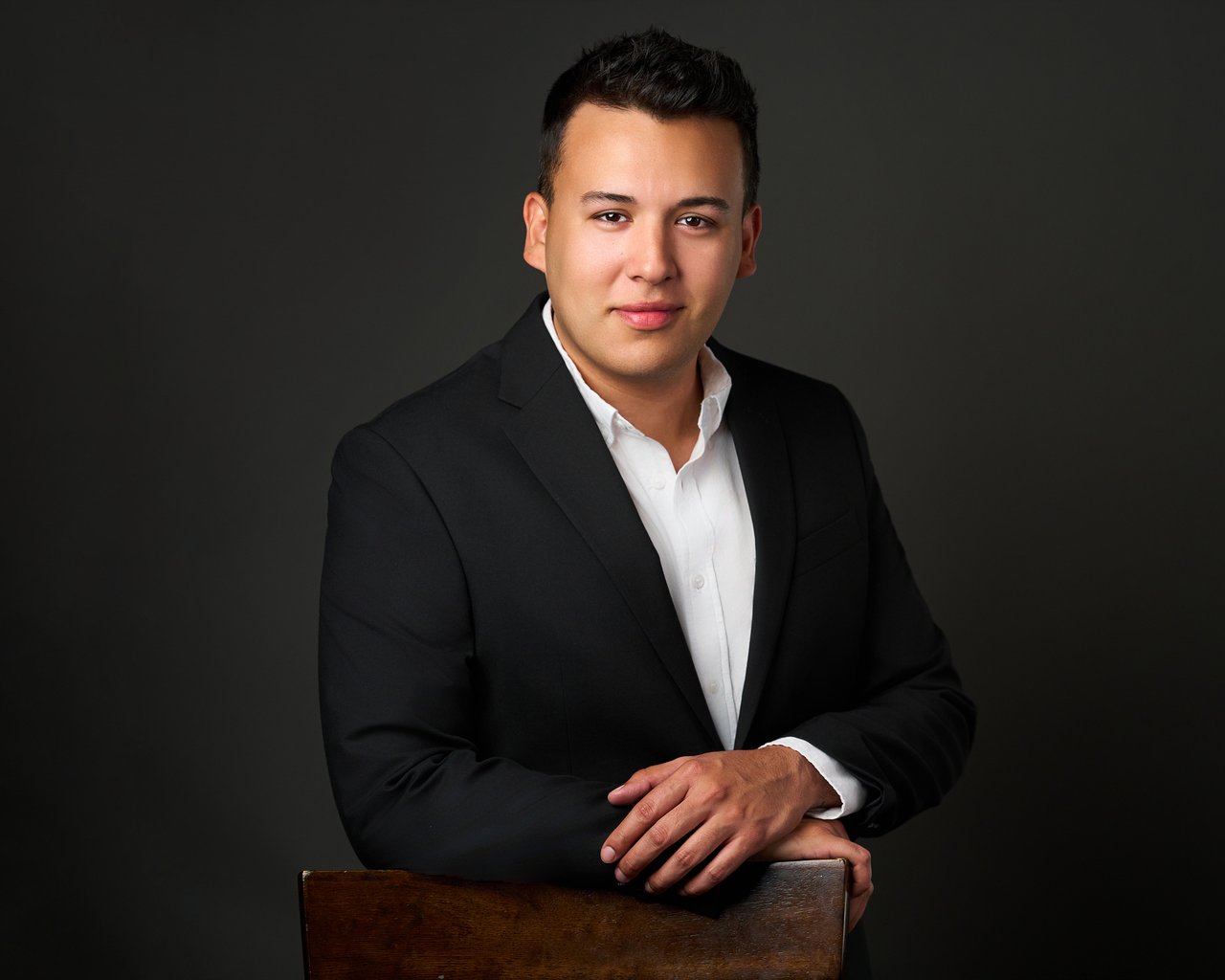 Robert Alexis Roberto in a black suit and white shirt, seated with his hands on a wooden chair.