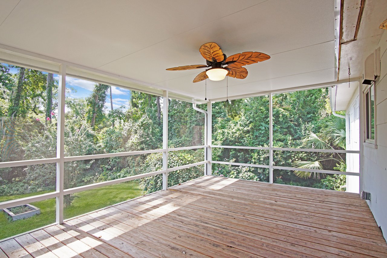 Spacious screened porch with wooden flooring, leafy ceiling fan, and lush green garden view. Bright, airy, inviting atmosphere, ideal for relaxation.