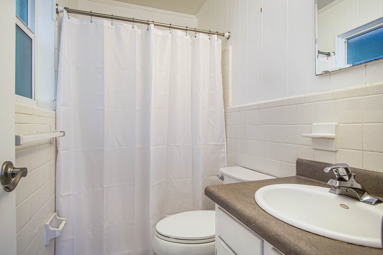 Compact bathroom with white tiles, a shower with a white curtain, a toilet, and a sink with a gray countertop. Bright, clean, and minimalistic.