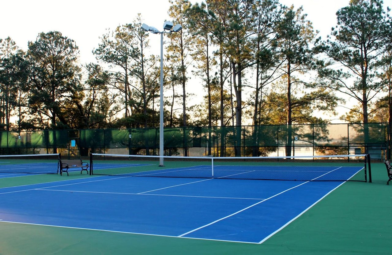 Outdoor tennis courts with blue playing surfaces, white nets, and tall pine trees beyond the fence.