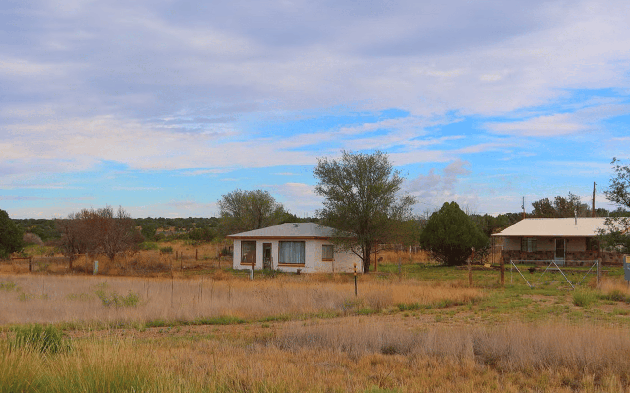 Rural property with two small buildings behind a metal gate, surrounded by dry grass and scattered trees.