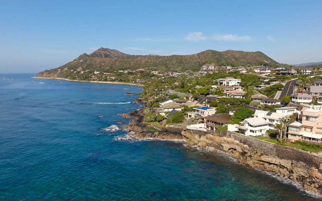 Kaimalino coastal neighborhood with homes along a rocky shore, turquoise water, and a mountain ridge.