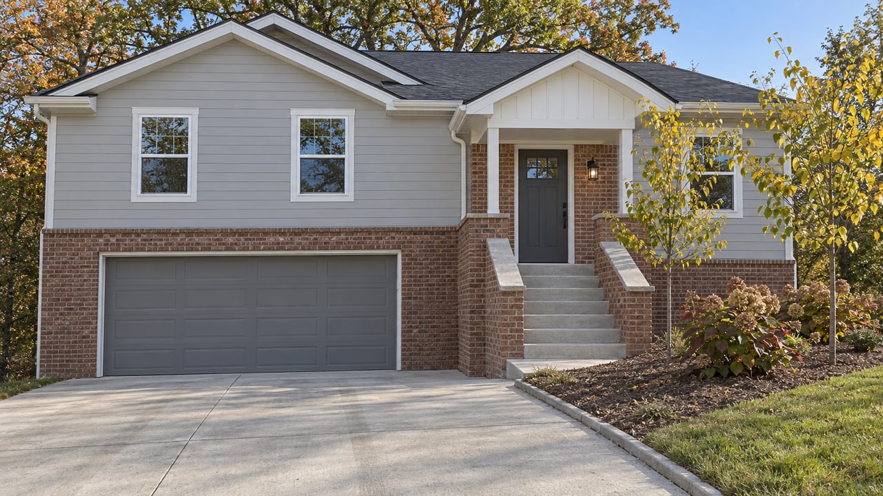 Raised-entry home with gray siding, brick lower walls, concrete driveway, and deciduous planting beginning to turn in autumn.