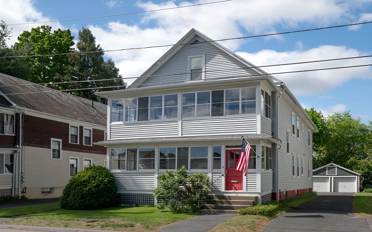 Gray multi-story home with an enclosed porch, red front door, and American flag.