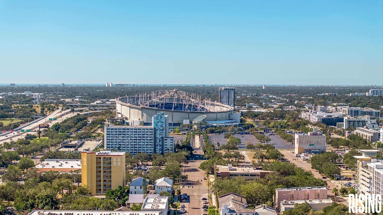 Aerial view of Tropicana Field with its roof structure exposed, surrounded by St. Petersburg buildings and roads.