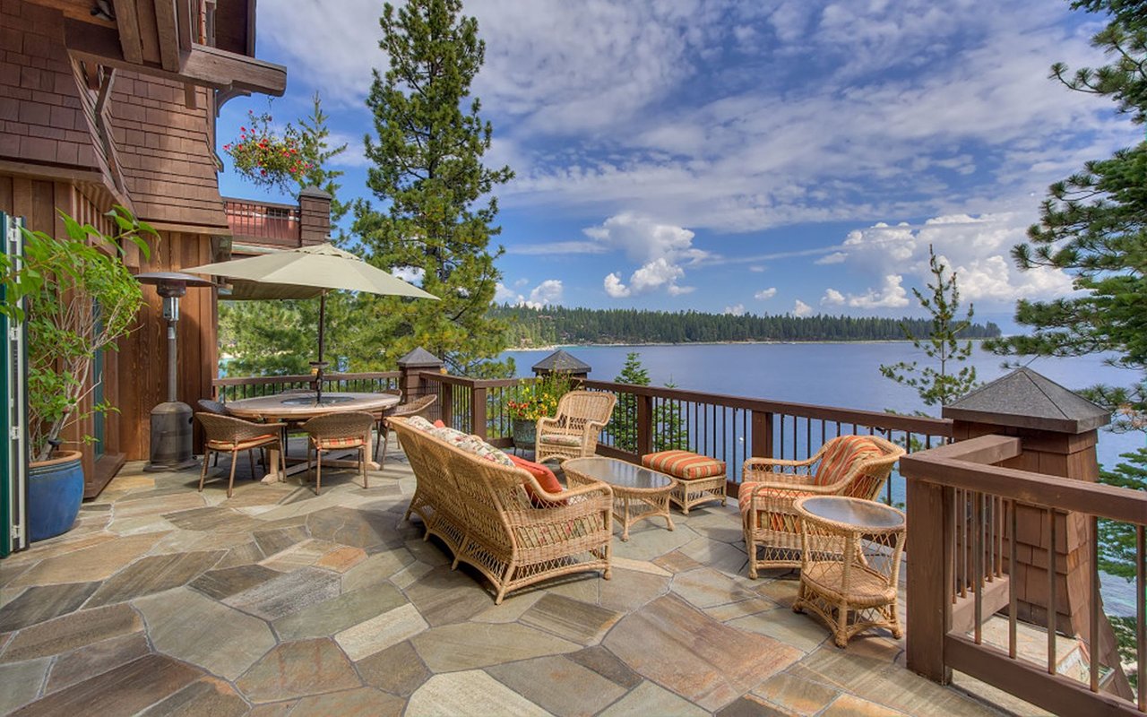 Furnished stone patio overlooking a lake, with wicker seating and a shaded dining area.