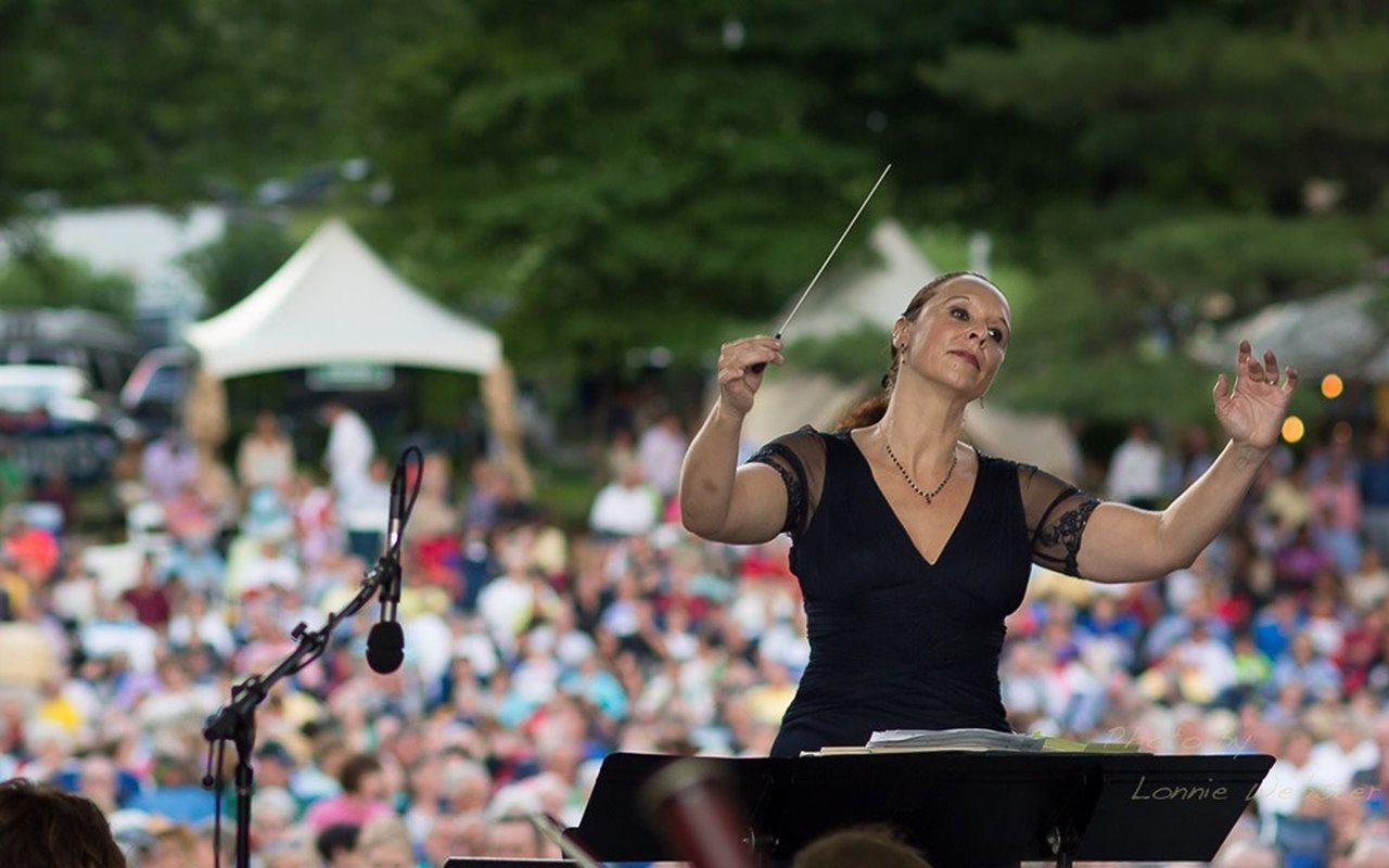 A woman conducts an outdoor concert before a large audience, raising her baton beside a microphone.