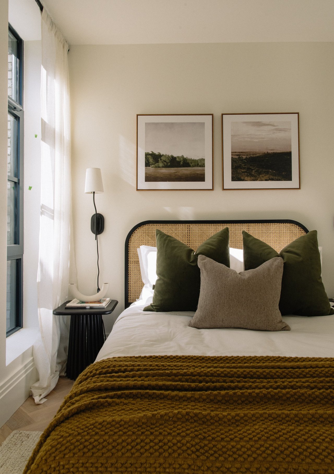 Bedroom with a cane headboard, olive pillows, landscape prints, and a chunky mustard-brown blanket.