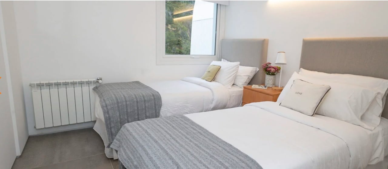 Bedroom with two white beds, gray headboards and throws, and a window above a radiator.